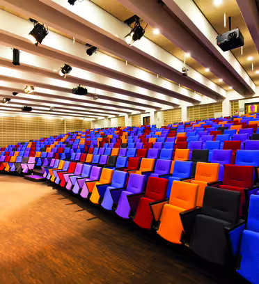 A modern lecture hall with a large array of colorful, tiered seating in shades of purple, red, orange, and gray. The room features a wooden floor, a podium at the front, and ceiling lights. The space is bright and well-organized.