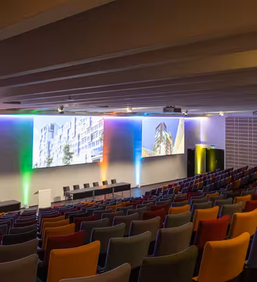 A modern auditorium with colorful seating and multiple large screens displaying architectural images on a stage, flanked by illuminated walls with rainbow lighting. The room is empty and well-lit, with a sleek design.