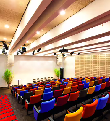 An auditorium with rows of colorful chairs in blue, orange, yellow, and red facing a stage. The ceiling features exposed beams with lights, and there are plants at the back corners. The walls have a modern, geometric design.