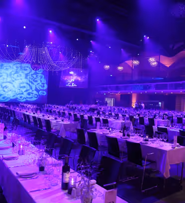 A large banquet hall lit with purple lights features long tables set with glasses and tableware. Elegant chandeliers hang from the ceiling, and ornate light patterns are projected on the walls.