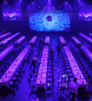 A dimly lit event hall is set with long rows of tables and chairs. Purple lighting creates a vibrant atmosphere, with a decorated stage at the front. Staff members are preparing the tables for an event.