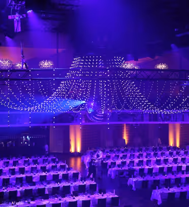 A large event venue is decorated with an elaborate ceiling of purple and white lights resembling a chandelier. Below are numerous tables covered with white tablecloths and black chairs, set for a formal gathering.