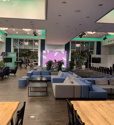 Spacious modern event hall with a large seating area featuring blue and gray sofas and chairs. The room is adorned with green plants and has a green and purple-lit stage at the center. Rows of chairs face the stage, and wooden tables are visible in the foreground.