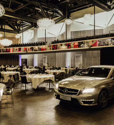 A silver luxury car is parked in an elegantly decorated banquet hall. The room features multiple round tables set with white tablecloths and chairs. Chandeliers hang from the ceiling, casting a warm glow.