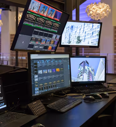 Control room with multiple screens displaying building images and graphics. A laptop shows TIVOLI Hotel & Congress Center on its screen. Modern design elements with hanging lights in the background.