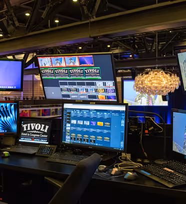 A tech control station with multiple computer monitors displaying various software and images. The room features chandeliers and illuminated seating in the background. The setup is housed in a modern venue, indicating an event or presentation.
