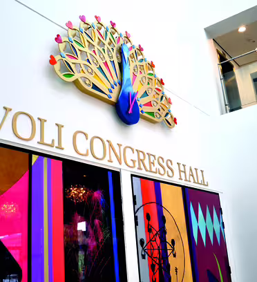 The image shows the entrance to Tivoli Congress Hall, featuring a colorful peacock emblem above the name in bold gold letters. Below are vibrant, geometric glass panels in various colors and patterns, filling the lower wall.