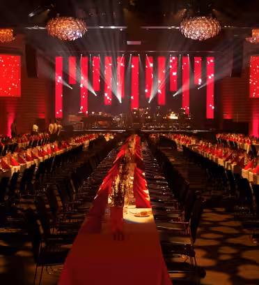 A grand banquet hall set for an event, featuring long tables adorned with red tablecloths and napkins. Chandeliers hang from the ceiling, and a stage with bright red lighting is visible at the front, creating an elegant ambiance.