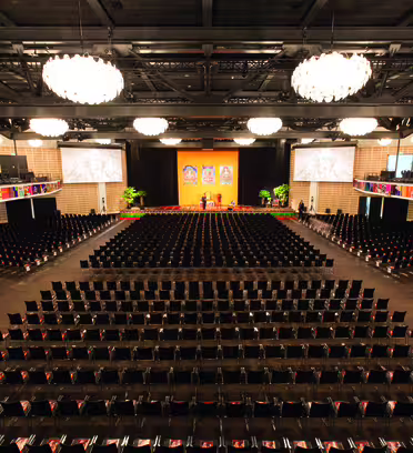 A large auditorium with rows of empty chairs facing a stage. The stage is adorned with colorful decorations and flanked by large screens. Chandeliers hang from the ceiling, and balconies line the sides. The atmosphere is formal and spacious.