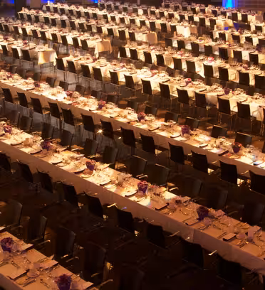 A large banquet hall with rows of parallel, elegantly set tables lined with white tablecloths, plates, and purple floral centerpieces. Black chairs are neatly arranged on both sides of each table. The room is well-lit, creating a warm ambiance.