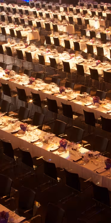 A large banquet hall with rows of parallel, elegantly set tables lined with white tablecloths, plates, and purple floral centerpieces. Black chairs are neatly arranged on both sides of each table. The room is well-lit, creating a warm ambiance.