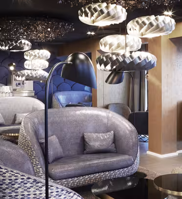 Modern lounge with curved, blue upholstered seating, circular floor lamps, and geometric ceiling lights. Dark blue, scalloped wall accents add texture. A reflective, dark glass coffee table is in the center, enhancing the contemporary ambiance.