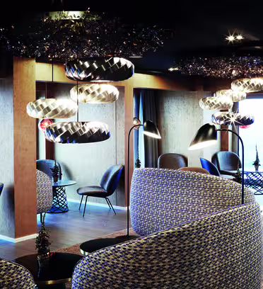 Modern lounge with patterned armchairs and sleek, metallic hanging lights. Tables with small decorations are placed between seating areas. The room features large windows and dark ceilings, creating a cozy and stylish atmosphere.